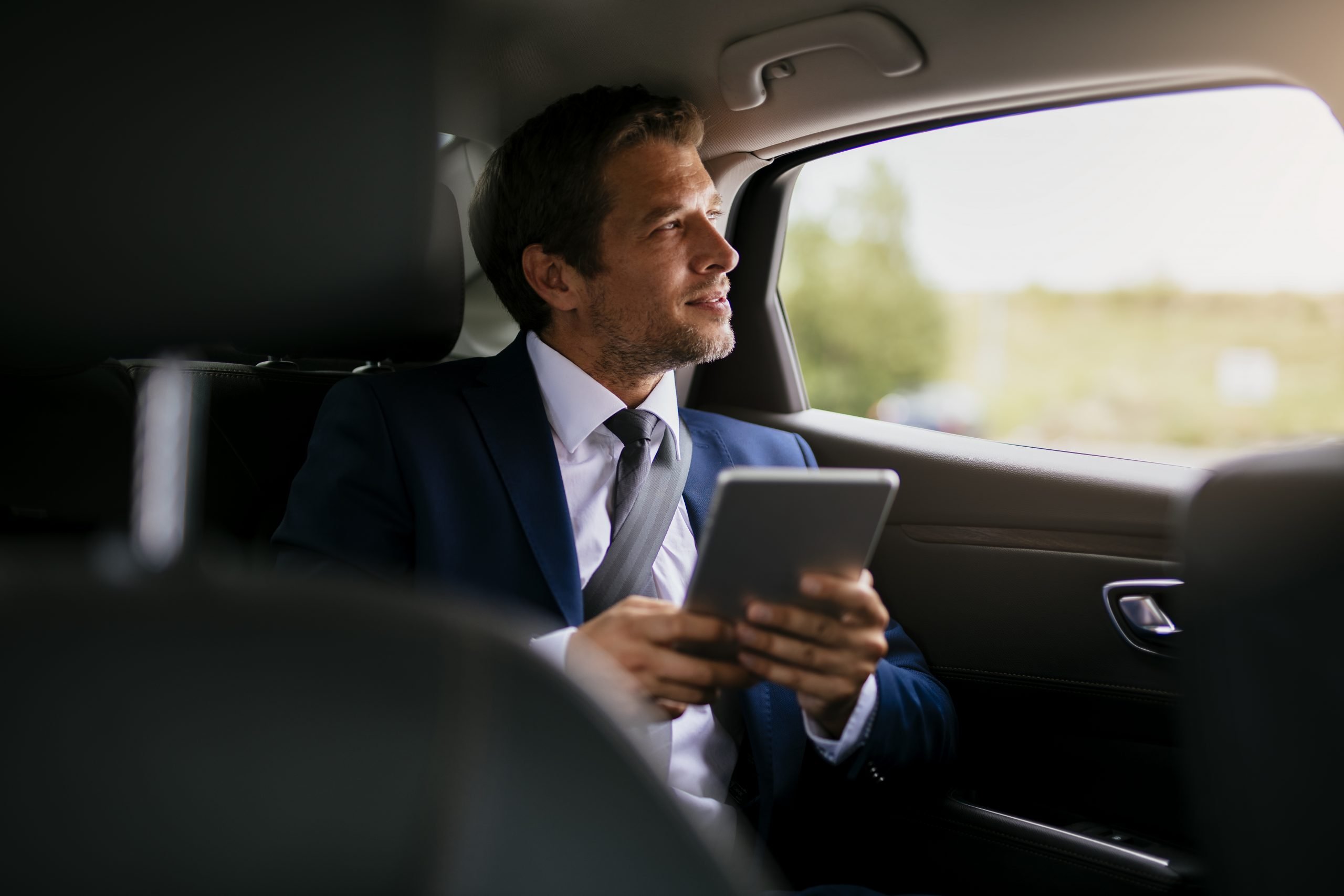 Handsome businessman sitting with digital tablet on the backseat of the car 