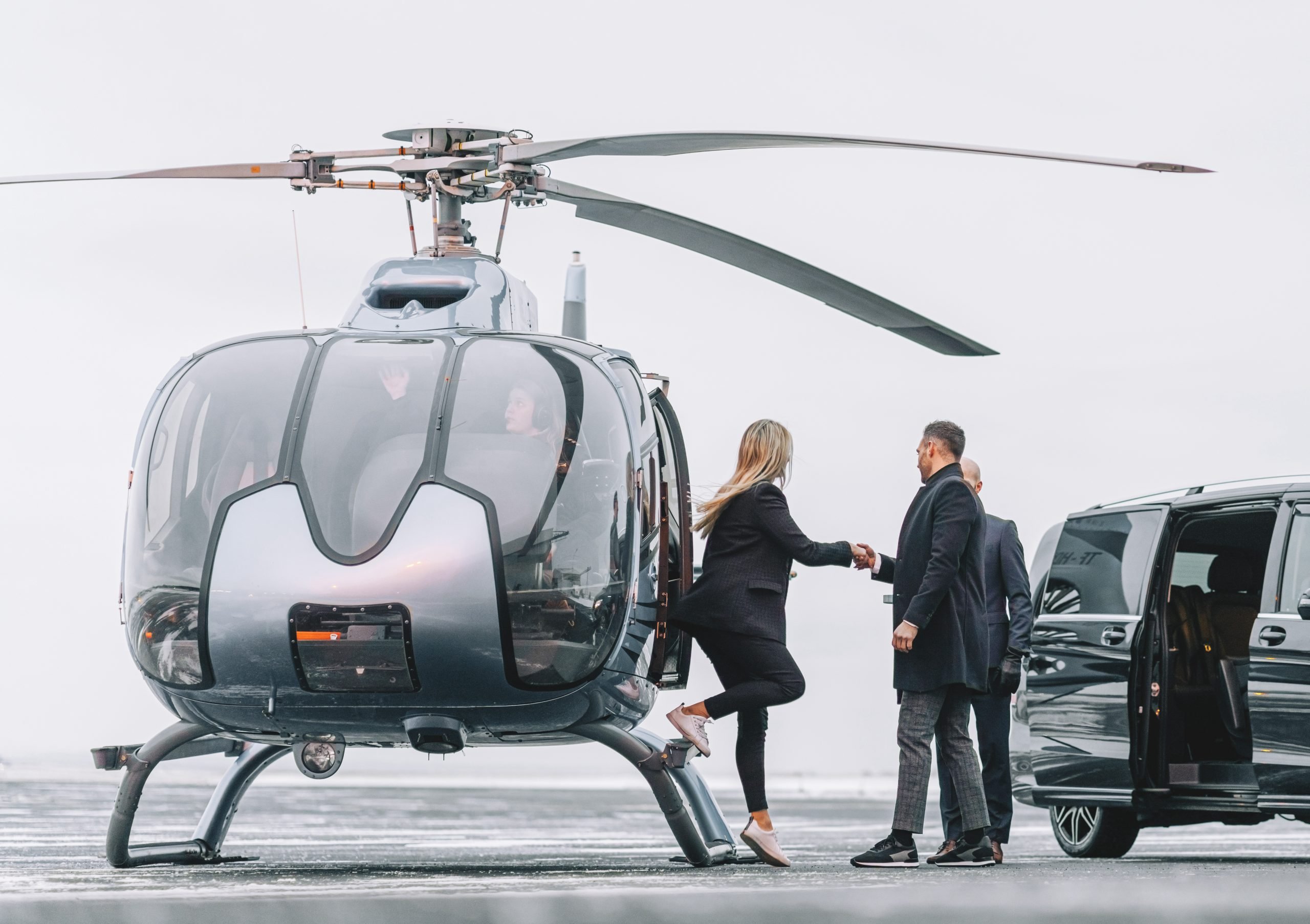 Woman getting down the helicopter with the help from a man with their chauffeur waiting by the car on the helipad. Couple disembarking their helicopter with chauffeur standing by.